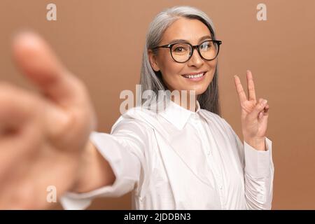 Lady lächelt und schaut isoliert auf beige auf die Kamera. Fröhliche ältere asiatische Frau, die Selfie macht, schöne reife Frau, die die Hand in der Siegesgeste hält Stockfoto