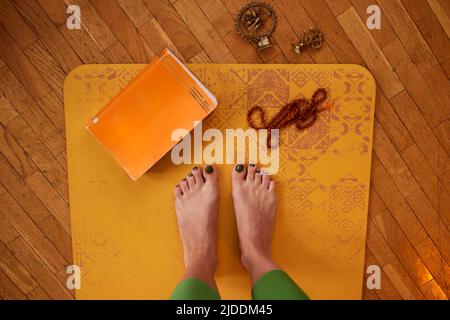 Draufsicht auf die Beine der Yogi-Frau auf der Matte mit Buch, Armband und Miniatur. Stockfoto
