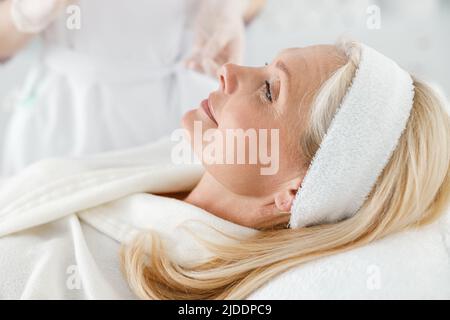 Schöne kaukasische blonde reife Frau mit geschlossenen Augen in Licht Kosmetologie Zentrum Stockfoto