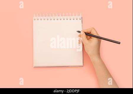 Mockup Notizblock. Frauenhand mit Bleistift neben einem leeren weißen Notizblock. Draufsicht der Hand, die den Bleistift gegen einen Spiralnotepad auf weißem Hintergrund hält Stockfoto