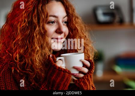 Ein süßes Ingwer-Mädchen mit lockigen Haaren sitzt morgens auf dem Stuhl zu Hause und trinkt ihren Kaffee. Stockfoto