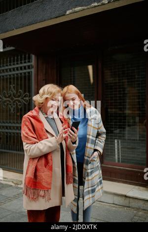 Eine Großmutter zeigt ihre heranwachsende Enkelin auf ihrem Telefon Bilder, während sie auf der Straße steht. Stockfoto