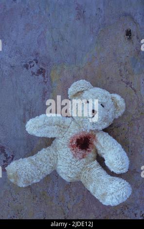 Blassbeige Teddybär liegt mit dem Gesicht nach oben auf violettem und orangefarbenem rauem Schiefer mit möglicher Schusswunde im Herzen Stockfoto
