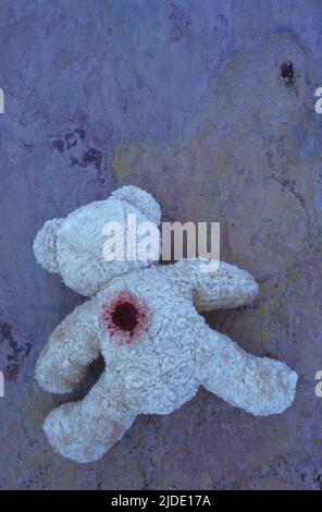 Blassbeiger Teddybär, der mit dem Gesicht nach unten auf violettem und orangefarbenem grobem Schiefer liegt, mit möglicher Schusswunde im Rücken Stockfoto