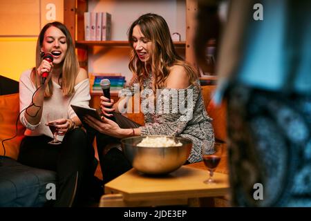 Mädchen sitzen zu Hause, trinken und singen ein Duett bei Karaoke-Abend. Stockfoto