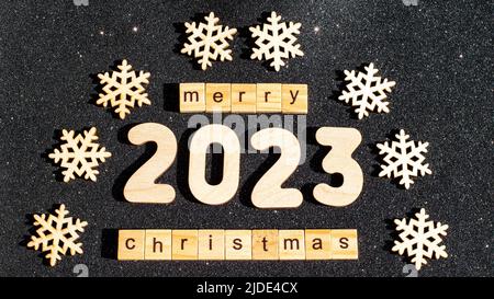 Konzept für Weihnachten und Neujahr - Holzschneeflocken Zahlen 2023 und die Inschrift HAPPY CHRISTMAS aus Holzbuchstaben auf schwarz glitzernden Sternen Stockfoto