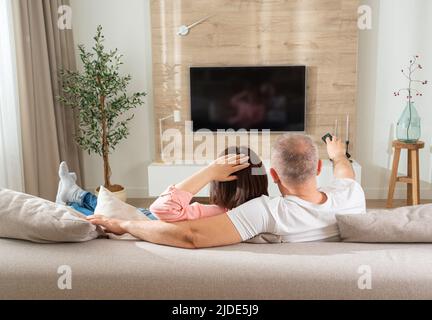 Entspanntes Paar schaltet den fernseher ein, während es auf dem Sofa im Wohnzimmer liegt Stockfoto