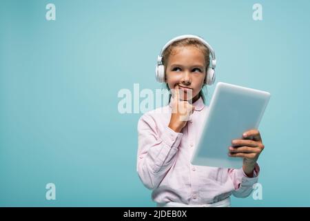 Nachdenkliche Schulkinder in Kopfhörern, die das digitale Tablet isoliert auf Blau halten Stockfoto