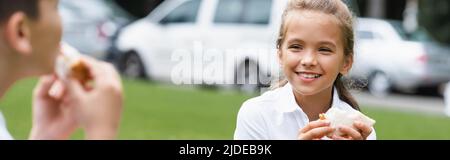 Positive Schülerin hält Sandwich in der Nähe verschwommener Freund im Freien, Banner Stockfoto