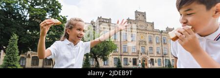 Fröhliches Schulkind hält Sandwich und winkt Hand in der Nähe von asiatischen firend im Park, Banner Stockfoto