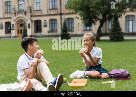Multiethnische Schüler halten Sandwiches in der Nähe von Rucksäcken auf Gras im Park Stockfoto