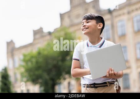 Blick aus dem niedrigen Winkel des lächelnden asiatischen Schuljungen, der im Freien einen Laptop hält Stockfoto