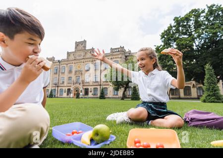 Schülerin hält Sandwich und winkt Hand in der Nähe von Lunchboxen und verschwommen asiatische Freundin auf Gras Stockfoto