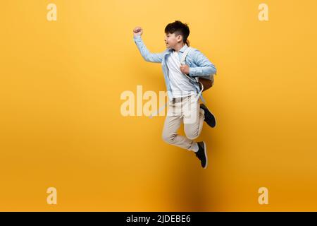 Seitenansicht eines asiatischen Schulkindes mit Rucksack, der auf gelbem Hintergrund springt Stockfoto