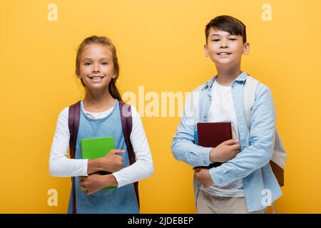Fröhliche multiethnische Jugendliche Schüler, die Bücher halten und die Kamera isoliert auf Gelb betrachten Stockfoto