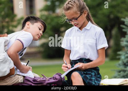 Multiethnische Schüler schreiben auf Notizbüchern auf der Bank im Park Stockfoto