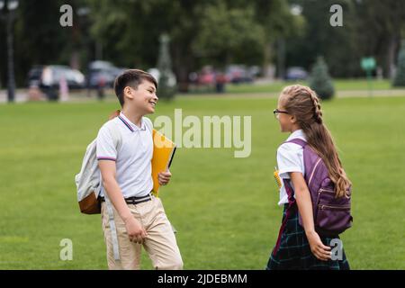 Lächelnde, multiethnische Schüler mit Notizbüchern, die im Park auf dem Rasen spazieren Stockfoto