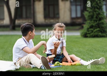 Positive Schulmädchen hält Sandwich in der Nähe asiatische Klassenkamerad auf Rasen im Park Stockfoto