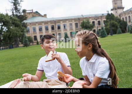 Lächelnder asiatischer Schuljunge hält Sandwich in der Nähe eines Freundes auf dem Rasen im Park Stockfoto