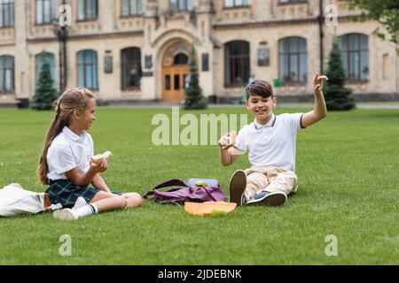 Asiatische Schuljunge hält Sandwich und zeigt mit dem Finger in der Nähe von Rucksack und Klassenkamerad im Park Stockfoto