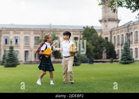 Positive multiethnische Kinder mit Rucksäcken und Notebooks, die im Freien auf Gras laufen Stockfoto