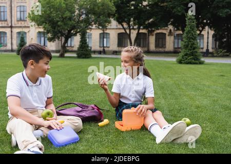 Aufgeregt Schulmädchen hält Sandwich in der Nähe von Lunchbox und asiatische Freundin mit Apfel auf dem Rasen Stockfoto