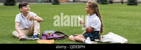 Schulmädchen hält Sandwich und spricht mit asiatischen Freund auf Gras im Park, Banner Stockfoto