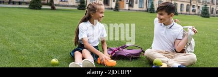 Lächelndes Schulmädchen hält Lunchbox in der Nähe einer asiatischen Freundin mit Rucksack auf Gras im Park, Banner Stockfoto