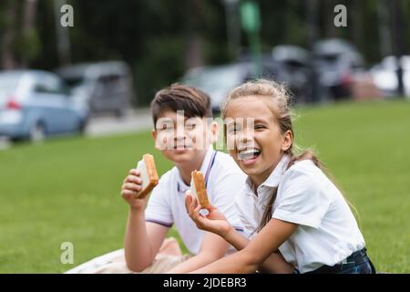 Aufgeregt Schulmädchen hält Sandwich in der Nähe verschwommen asiatische Klassenkamerad im Park Stockfoto