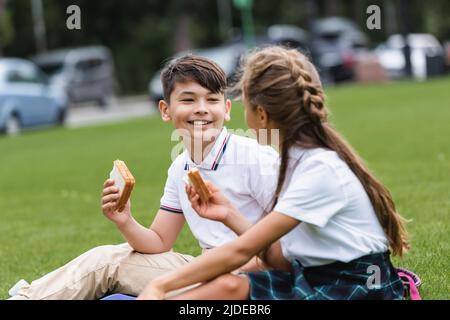 Lächelnder asiatischer Schuljunge hält Sandwich in der Nähe verschwommener Freund auf dem Rasen im Park Stockfoto