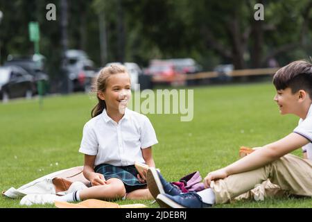 Glücklich Schulmädchen hält Sandwich in der Nähe verschwommen asiatische Klassenkamerad auf Gras im Park sitzen Stockfoto