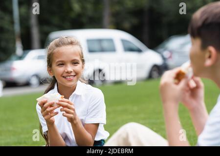 Lächelndes Schulmädchen hält Sandwich in der Nähe verschwommener Klassenkameradin im Park Stockfoto