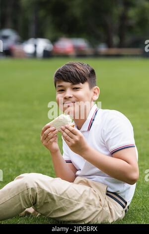 Lächelnder asiatischer Schuljunge hält Sandwich auf Gras im Park Stockfoto