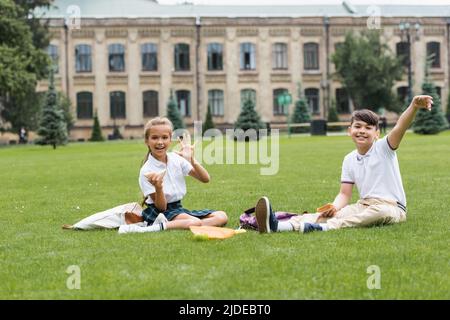 Positive Schülerin hält Sandwich und winkt Hand an der Kamera in der Nähe von asiatischen Freund auf Rasen im Park Stockfoto