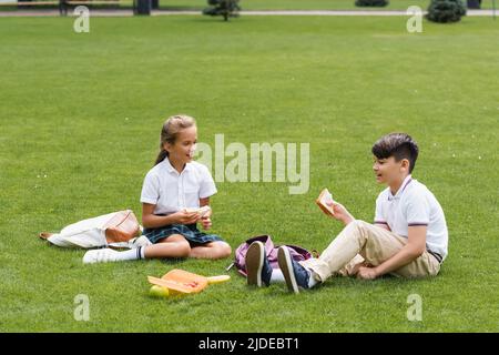 Multiethnische Schüler halten Sandwiches in der Nähe von Rucksäcken auf Gras im Park Stockfoto