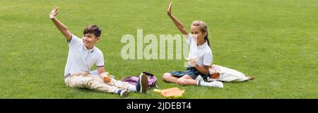 Positive multiethnische Schüler halten Sandwiches in der Nähe von Rucksäcken auf Gras im Park, Banner Stockfoto