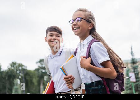 Positive Schüler mit Rucksack und Notebooks gehen in der Nähe verschwommener asiatischer Freund im Freien Stockfoto