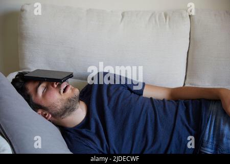 Der junge Mann schläft mit Ebook auf dem Kopf auf dem Sofa Stockfoto
