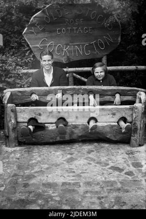 1935, historisch, ein Vater und eine Tochter, die für ihr Foto in „Ye Olde Stocks“ im Rose Cottage in Cockington, Torquay, Devon, England, Großbritannien, sitzen. Vorräte waren Holzgeräte, die als eine Form der "Bestrafung" und "Scham" für lokale Straftäter von Kleinkriminalität verwendet wurden und die Straftäter mit Füßen und Händen fesselten. Auf Dorfgrüns in Großbritannien gesehen, waren sie bis zum Ende des 18.. Jahrhunderts im Einsatz. Stockfoto