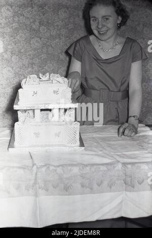 1950s, historisch, eine junge Frau, die ein Partykleid, Perlenohrringe, eine Halskette und ein großes Lächeln trägt und neben ihrem besonderen Geburtstagstorte aus dem Jahr 21. steht, Stockport, England, Großbritannien. Stockfoto