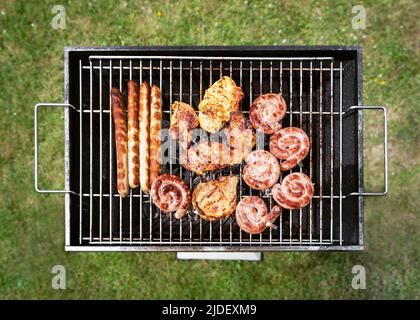 Leckere Auswahl an Fleisch auf einem Sommergrill mit Steaks, Huhn, Würstchen, Spiralen Rinderwurst. Blick von oben auf grünes Gras. Stockfoto