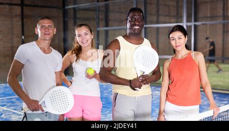 Lächelnde Männer und Frauen mit Schlägern und Bällen, die auf dem Padel-Platz in der Halle posieren Stockfoto