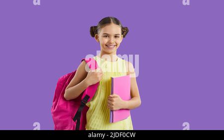 Glückliches Schulmädchen, das auf purpurem Hintergrund steht, pinke Tasche und Bücher hält und lächelt Stockfoto