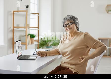 Glückliche ältere Frau, die zu Hause am Schreibtisch sitzt und einen Laptop für Fernarbeiten oder Studieren benutzt Stockfoto