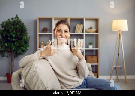 Glückliche junge Frau, die zu Hause auf dem Sofa sitzt, lächelt und zeigt die Daumen nach oben Stockfoto