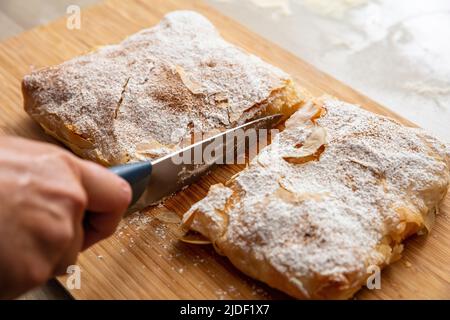 Griechisches Gebäck mit Sahnezucker-Pulver und gemahlenem Zimt. Hand mit einem Messer schneiden Bougaza, traditionelle Pudding Pie auf einem Holzbrett, Nahaufnahme Stockfoto