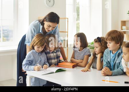 Die Lehrerin hilft ihren glücklichen kleinen Schülern, die in ihren Notizbüchern schreiben Stockfoto