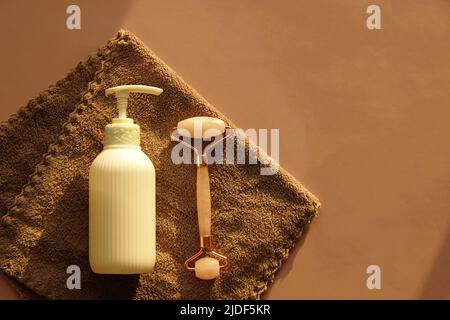 Set für die Gesichtspflege zu Hause auf braunem Handtuch. Gesichtsbehandlung Rose Quarz Massage Roller und сosmetic Pumpe Flasche feuchtigkeitsspendende Lotion für die Pflege.Kosmetische Lifting-Kit Stockfoto