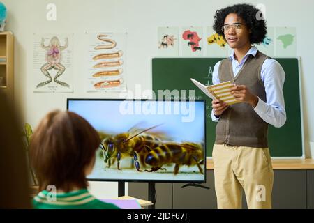 Afroamerikanischer Kerl mit Copybook, das an interaktiver Ausstellung mit Bienen steht, während er eine Präsentation vor Klassenkameraden während des Unterrichts macht Stockfoto