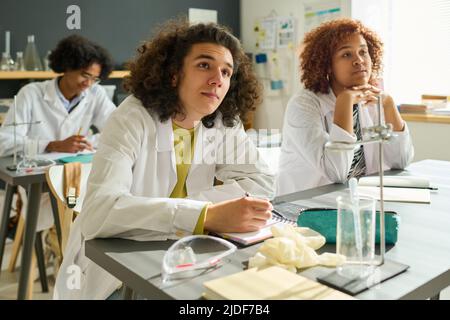 Gruppe von cleveren interkulturellen Schülern der Highschool in Laborkittel, die der Erklärung ihres Lehrers lauschen, während sie am Schreibtisch bei der Chemie sitzen Stockfoto
